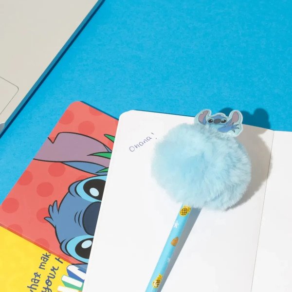 Stylo Disney Stitch Tropical avec pompon bleu posé sur un carnet, avec visuel Stitch en arrière-plan.