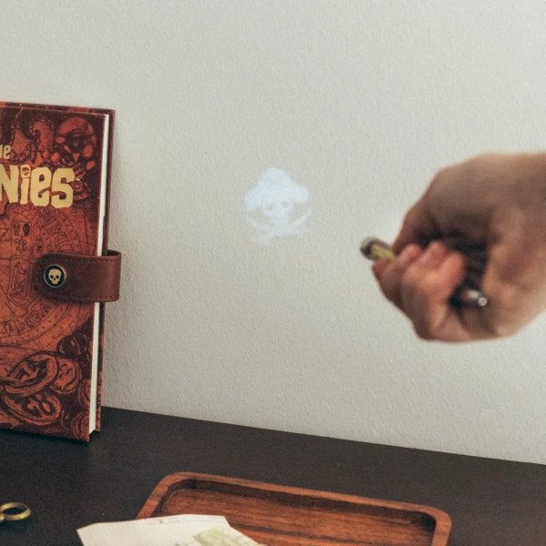 Stylo projecteur Les Goonies projetant un symbole de pirate à côté du carnet.