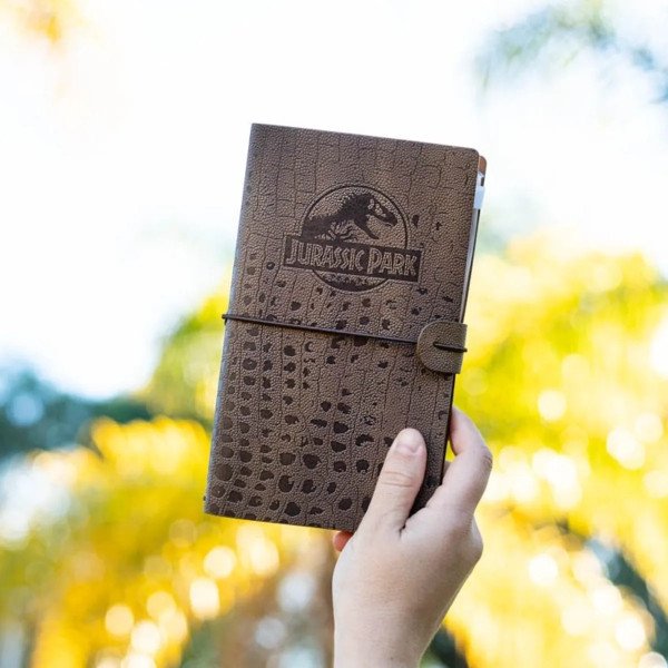 Carnet de voyage Jurassic Park tenu à la main en extérieur, couverture fermée avec logo embossé.