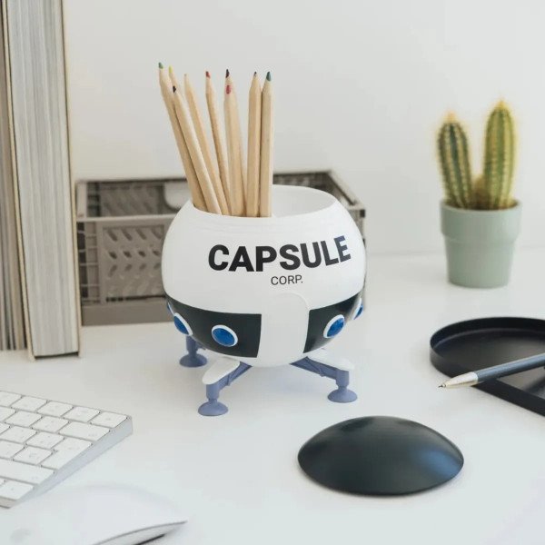 Pot à crayons Dragon Ball Z Capsule Corp posé sur un bureau, rempli de crayons avec couvercle à côté.
